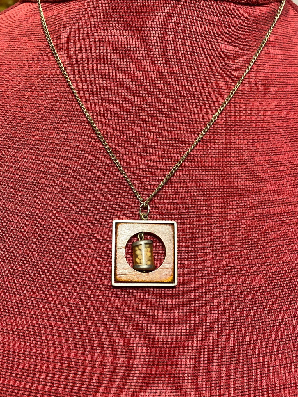 Vintage necklace with pendant containing small mustard seed bottle inside a circular opening inside a piece of wood encased in a metal frame hanging on a 19 inch chain